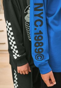 Chemise noire à manches longues avec motif à carreaux blancs sur la manche ; chemise bleue à manches longues avec un grand texte noir "NYC.1989©" sur le bras.