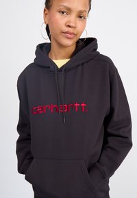 Black hoodie with front pocket, featuring red embroidered "carhartt" logo. Made of soft fabric, with a drawstring hood and ribbed cuffs.