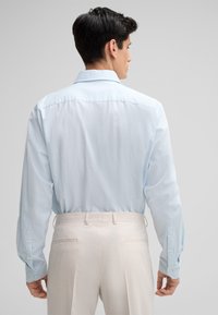 Hellblaues Button-up-Hemd mit langen Ärmeln, Rückansicht, kombiniert mit hellbeigen Hosen. Glatte Textur, klassischer Kragen und verstellbare Bündchen.