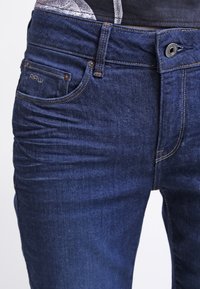 Tmavě modré džínové kalhoty s hladkou texturou, vybavené poutky na pás, předními kapsami a malým logem na pásku.