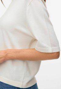 Hvid kortærmet top med rund halsudskæring og ribbet tekstur. Har en kontrasterende kant og manchetter, parret med blå jeans.