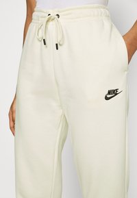 Calças de moletão cor de creme claro feitas de um tecido suave, com cintura elástica ajustável e um logotipo preto da Nike na perna esquerda. Textura suave.