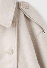 Gros plan d'un manteau en laine beige montrant de gros boutons, le col et le détail de l'épaulette d'épaule.