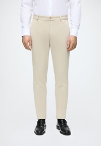 Smėlio spalvos kelnės, pagamintos iš lygaus audinio, turinčios prigludusį dizainą, šonines kišenes ir užsegimą vienu mygtuku, derinamos su juodais loafers.