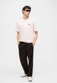Άνδρας που φορά γυαλιά ηλίου, ανοιχτό ροζ T-shirt Adidas με λογότυπο, χαλαρό μαύρο παντελόνι με διακριτικό σχέδιο και λευκά παπούτσια Adidas, στέκεται με τα χέρια στις τσέπες.