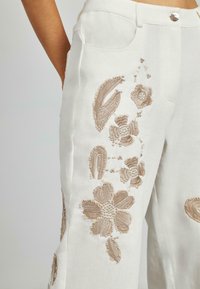Pantaloni in denim bianco ricamati con motivi floreali e fogliari beige, dotati di tasche e chiusura frontale con bottone. Tessuto morbido e vestibilità su misura.