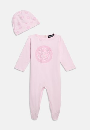 Zachtroze babyrompertje met Medusa-logo op de borst en bijpassend hoedje met cirkelpatroon, beide gemaakt van gladde stof.