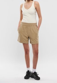 Lightweight beige shorts with an elastic waistband, side pockets, and a soft texture. Paired with a white ribbed tank top and black sneakers.