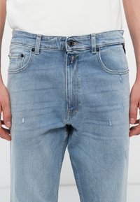 Blugi din denim de culoare albastru deschis, cu închidere prin buton și fermoar, cinci buzunare și detalii subtile de destrămare pe țesătură.