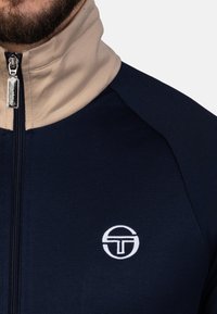 Marineblaue und beige Zip-Jacke mit hohem Kragen, strukturiertem Stoff und einem weißen Logo auf der Brust. Detaillierte Nähte an den Schultern.