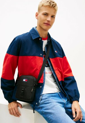 Ung mand iført en marineblå og rød jakke med en hvid grafisk t-shirt og blå jeans, bærende en sort skuldertaske med logo.