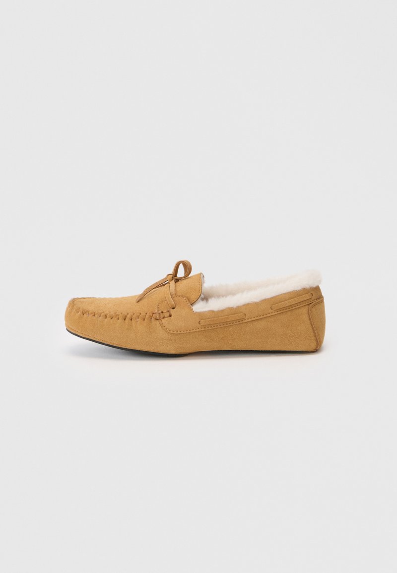 Suede moccasin-sandaalit vaaleassa ruskeassa sävyssä, joissa on valkoinen fleecepäällyste, ommellut yksityiskohdat ja nauhakiinnitys, esitetty sivuprofiilissa valkoista taustaa vasten.