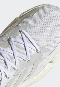 Baskets blanches de sport avec une tige en mesh texturé, des lacets fins blancs et des accents en caoutchouc gris clair le long de la semelle intermédiaire et des côtés.