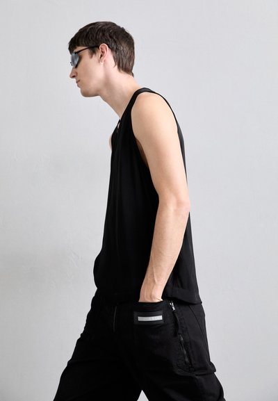 Top negru fără mâneci, fabricat dintr-un material neted, combinat cu pantaloni cargo negri care au un buzunar cu fermoar și un patch cu logo pe spate.