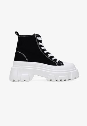 Zapatillas altas de lona negras con cordones blancos, suela blanca gruesa, detalles cosidos y un patrón de tacos distintivo en la suela exterior.