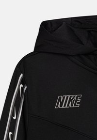 Svart Nike-hoodie med hög krage, snedställda sömmar och en vit logga. Sidodetaljer visar upprepade små Nike-swooshes.