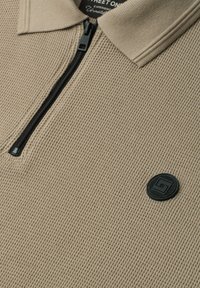 Polo en maille beige avec fermeture éclair noire à l’avant et écusson rond noir avec logo sur la poitrine. Col côtelé et tissu texturé.