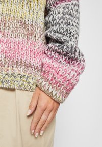 Hand rustend op beige broek, gekleed in een veelkleurige dikke gebreide trui met roze, geel, grijs en witte draad in een textuurpatroon.