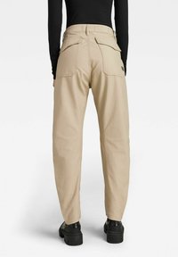 Osoba v béžových kalhotách s vysokým pasem a velkými kapsami na zadní straně, s černým topem s dlouhým rukávem a robustními černými platformovými botami, stojící čelem odvrácena.