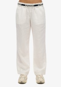 Witte linnen broek met een zwarte tailleband met het "SUPERDRY" logo. Ze hebben een relaxed fit en verticale textuurdetails. Gecombineerd met beige sneakers.