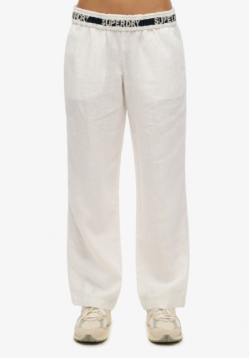 Witte linnen broek met een zwarte tailleband met het "SUPERDRY" logo. Ze hebben een relaxed fit en verticale textuurdetails. Gecombineerd met beige sneakers.