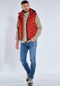 Rote Steppweste mit hohem Kragen, Reißverschluss vorne und Taschen, darüber ein beiges Hemd mit Knöpfen und blaue Jeans.