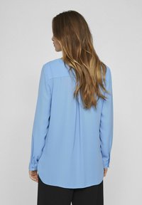 Langermet blå bluse i glatt stoff, med rund nederkant, knappede mansjetter og en avslappet passform. Bakside viser skulderplissédetalj.
