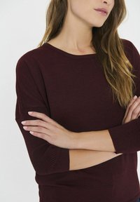 Burgundska pulover srajca iz mehkega rebrastega materiala z oblikovanim izrezom in rokavi do tridesetih, ki ima sproščen kroj.
