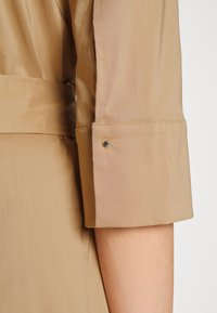 Manche de veste couleur beige avec une texture lisse, un revers roulé et un détail de bouton avec une tige métallique contrastante.