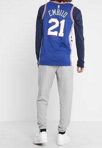 Persona che indossa una maglia da basket blu "Embiid 21", maniche lunghe blu scuro, pantaloni grigio chiaro e sneakers bianche, in piedi su uno sfondo semplice.