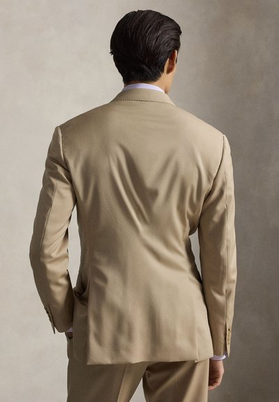 Uomo in piedi di spalle alla macchina fotografica, indossa una giacca da completo beige e pantaloni abbinati su uno sfondo uniforme.