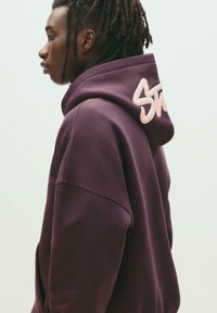 Felpa viola con un grande logo rosa "Stussy" ricamato sulla schiena. Tessuto morbido, vestibilità comoda, con cappuccio regolabile.