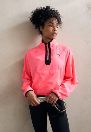 Cazadora de pullover de color rosa brillante con media cremallera y detalles en negro, que presenta un bolsillo frontal y puños elásticos, combinada con leggins negros.
