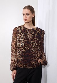 Blouse en tissu transparent à imprimé léopard, avec un col rond et une ouverture en clé à l'avant ; manches longues avec poignets élastiques. Tons foncés sur fond beige.