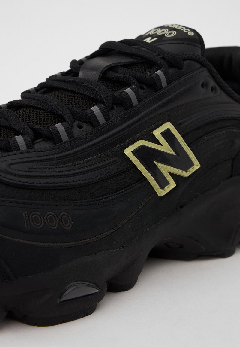 Sneaker nera con materiali in rete e gomma, caratterizzata da un prominente logo "N" nero e giallo, superficie testurizzata e lacci visibili.
