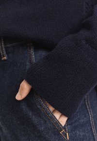 Pulover tricotat navy cu manșete cu nervuri, acoperind parțial o mână. Asortat cu blugi din denim închis, cu cusături în contrast.