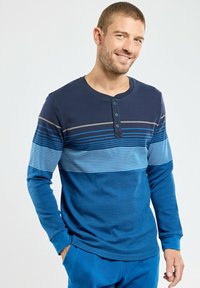 Homme souriant portant une chemise henley à manches longues à rayures bleues avec trois boutons, se tenant avec une main dans la poche devant un fond uni.