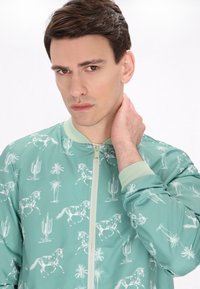 Lichtgroene jas met een patroon van een paard en cactussen, geribde manchetten, een rits aan de voorkant en een gladde textuur.