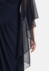 Abito blu navy con sovrapposizione trasparente in filo metallico nero. La sovrapposizione presenta un bordo ondulato ed è leggera, aggiungendo texture e movimento.