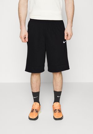 Mann trägt schwarze Nike-Shorts, schwarze Nike-Socken und leuchtend orangefarbene Sportschuhe, steht vor einem schlichten weißen Hintergrund.
