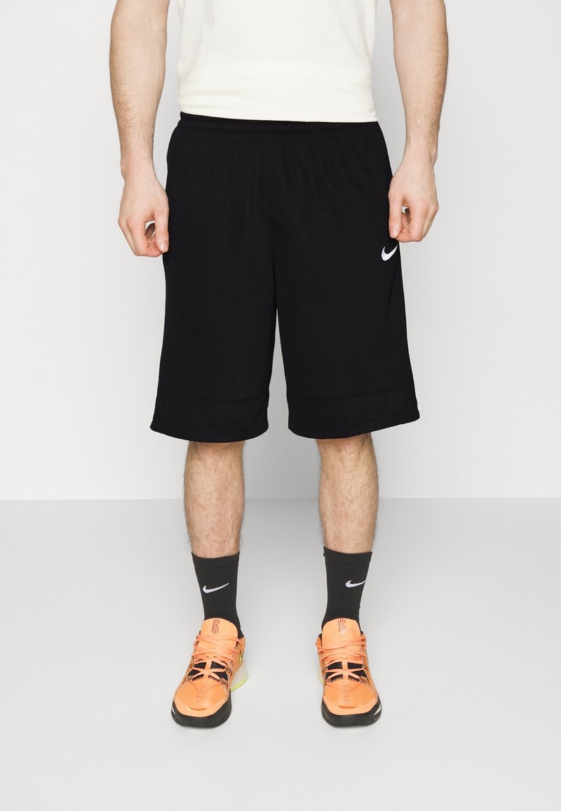 Homme portant un short Nike noir, des chaussettes Nike noires et des chaussures de sport orange vif, debout devant un fond blanc uni.
