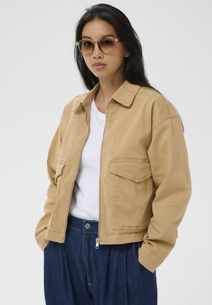 Smėlio spalvos drobinė striukė su apykakle ir priekinėmis kišenėmis, derinama su balta palaidine ir mėlynomis aukšto liemens kelnėmis. Išbaigimą suteikia skaidrūs akiniai nuo saulės.