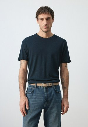 Uomo con capelli castani corti che indossa una maglietta blu scuro, jeans blu, una cintura beige e mostra tatuaggi su entrambi gli avambracci.