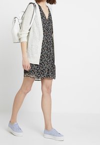 Žena v černých květinových šatech, bílé pletené cardigan, levandulových teniskách a s bílým batohem, kráčející proti bílému pozadí.