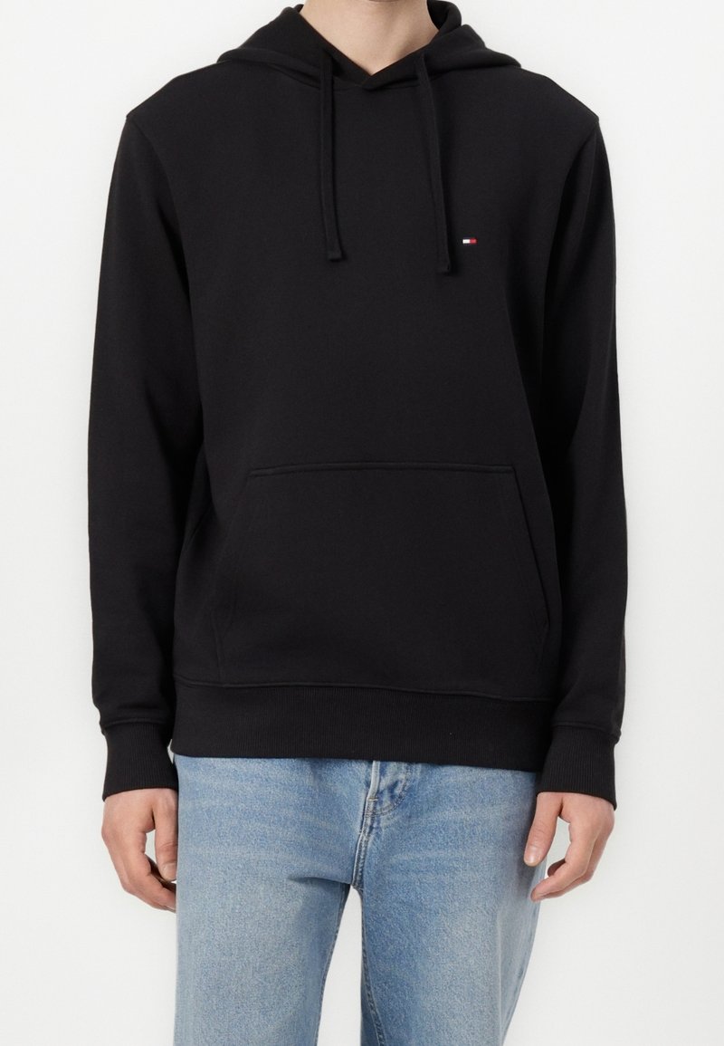 Homme portant un sweat à capuche noir avec une poche avant et un petit logo, associé à un jean bleu clair, debout devant un fond blanc.
