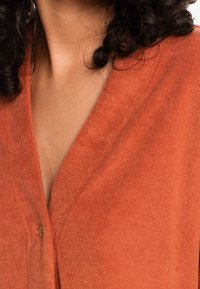 Kardigan w kolorze rdzy z dekoltem w kształcie litery V, wykonany z miękkiego materiału. Posiada jeden guzik przy dekolcie. Gładka faktura.
