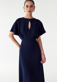 Laivansiniset mekko, jossa on lyhyet, liehuvat hihat ja avokerho kaula-aukko. Sileä pinta ja istuva siluetti, joka ulottuu polven alapuolelle.