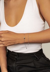 Silver bracelet with heart charm, featuring a delicate chain and bead accents. Worn on the wrist against a white top and black pants.