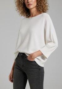 Weißer Strickpullover mit lockerem Schnitt, gerippten Bündchen und rundem Ausschnitt, kombiniert mit dunkelgrauen Jeans. Leichtes Material und lässiges Design.