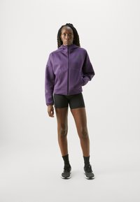 Lila zip-jacka med hög krage, med en slät textur och subtilt logodetalj. Matchas med svarta shorts och sportskor.
